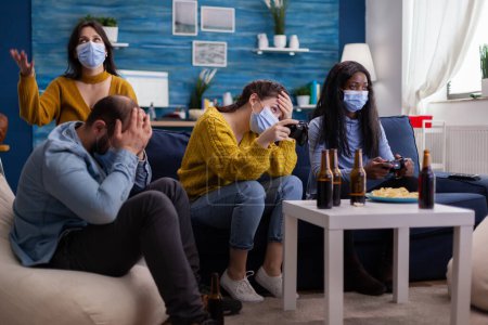 Photo for Disappointed diverse people loosing video games at home respecting social distancing because of corona outbreak wearing face mask against spreading virus. New normal party social distance - Royalty Free Image