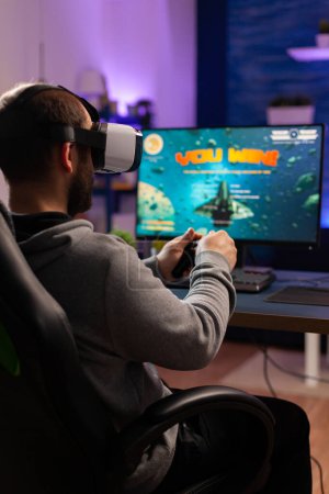 Téléchargez les photos : Gamer perdre jeu de tournoi de tir de l'espace en ligne portant casque de réalité virtuelle. Esport homme assis sur chiar de jeu tard dans la nuit dans le home studio en utilisant un équipement professionnel - en image libre de droit