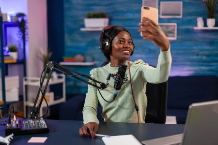 Photo for Afro woman taking selfie with smartphone and using professional gear to record episode in living room. On-air online production internet podcast show host streaming live content, recording digital - Royalty Free Image