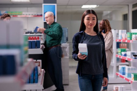 Photo for Asian client holding drugs package reading presription before buy it. Woman customer walking between shelves shopping for supplements, vitamin, medication in pharmaceutical shop. Health care products - Royalty Free Image