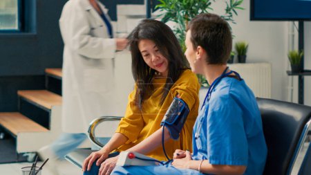 Foto de Enfermera haciendo consulta de cardiología con paciente asiática en vestíbulo de sala de espera, midiendo hipertensión con tonómetro. Comprobación de la presión arterial y del pulso con instrumento médico. - Imagen libre de derechos