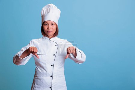 Photo for Senior asian cook wearing kitechen uniform doing dislike sign after looking at culinary dinner meal. Cheerful woman chef showing thumbs down gesture, preparing gastronomy healthy dish - Royalty Free Image