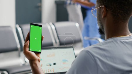 Photo for Male patient using greenscreen template on smartphone while he waits in facility waiting area. Young man looking at isolated mockup display and chroma key, blank copyspace on mobile phone screen. - Royalty Free Image