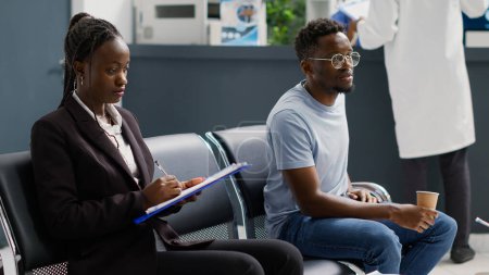 Téléchargez les photos : Les Afro-Américains assis dans la salle d'attente à la réception clinique, attendant de commencer un rendez-vous de contrôle médical. Lobbying des personnes en établissement ayant une consultation ou un examen. - en image libre de droit