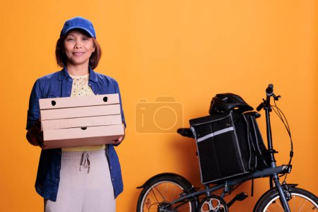 Téléchargez les photos : Travailleur du restaurant portant uniforme bleu tenant pile de pizza livrer au client sur le vélo. Un livreur de nourriture debout dans un studio avec un fond jaune. Service alimentaire et transport - en image libre de droit