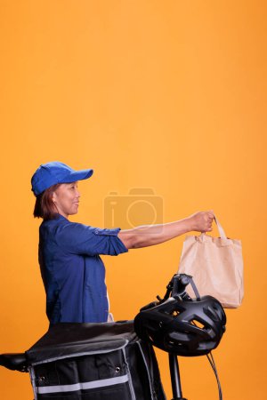 Téléchargez les photos : Un livreur de plats à emporter âgé livre des aliments à emporter dans des sacs en papier recyclés au client, en utilisant le vélo comme moyen de transport. Employé du restaurant portant t-shirt bleu et casquette tout en travaillant comme messager - en image libre de droit