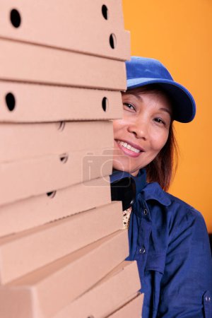 Photo for Smiling asian senior delivery employee holding pizza boxes, delivering to client during lunch time. Pizzeria worker hidden behind a large stack of takeout food order. Take out food concept - Royalty Free Image