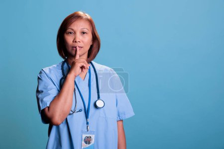 Photo for Senior nurse showing shhh taboo sign with finger to lips while working at medical expertise during checkup visit appointment. Specialist assistant wearing blue uniform and stethoscope. - Royalty Free Image