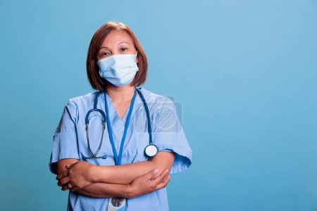 Téléchargez les photos : Infirmière asiatique debout en studio avec le bras croisé sur fond bleu lors de la visite de contrôle médical rendez-vous. Assistant principal portant un masque protecteur contre le coronavirus, système de soins de santé - en image libre de droit