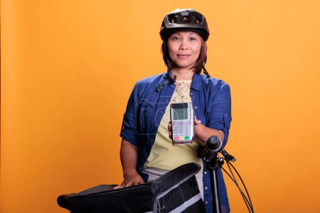 Photo for Portrait of deliverywoman with blue uniform and helmet delivering fast food order giving POS terminal to customer. Pizzeria employee using bike as transportation while carrying takeout food backpack - Royalty Free Image