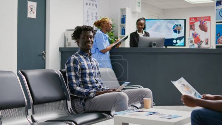 Téléchargez les photos : Afro-Américain en attente d'assister à la consultation avec le médecin, assis à la réception de l'hôpital. Jeune adulte en salle d'attente ayant rendez-vous d'examen avec un spécialiste pour guérir la maladie au centre de santé. - en image libre de droit