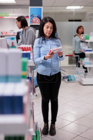 Téléchargez les photos : Client asiatique lecture de pilules boîte dépliant avant d'acheter des produits de santé à partir des étagères de la pharmacie. Client de pharmacie regardant les emballages de médicaments et de vitamines, les médicaments de cardiologie. - en image libre de droit