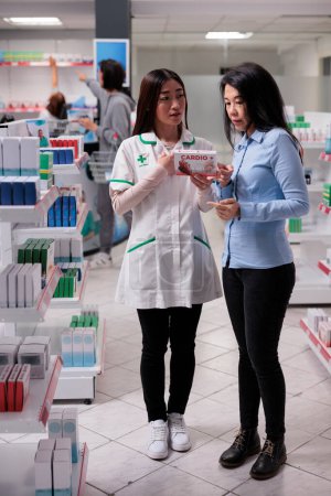 Téléchargez les photos : Assistant asiatique et client examinant la boîte de pilules de cardiologie des étagères de pharmacie, l'achat de suppléments médicinaux en pharmacie. Client parlant de fournitures pharmaceutiques. - en image libre de droit