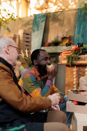 Téléchargez les photos : Jeune Afro-Américain mangeant des pommes alors qu'il était assis à table avec un vendeur amical près du stand avec des produits biologiques, homme noir dégustant des fruits et légumes frais des agriculteurs locaux à la foire d'automne - en image libre de droit