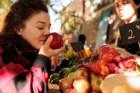 Healthy customer enjoying organic natural smell of apples, standing in front of farmers market stand. Woman smelling bio fruits before buying homegrown eco produce from counter.