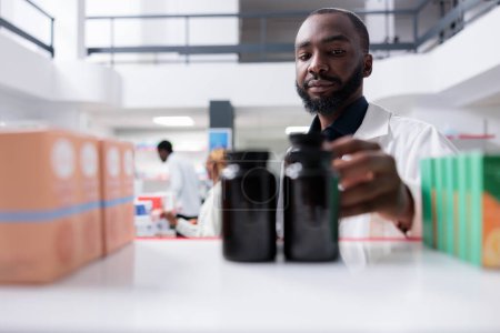 Photo for African american pharmacist taking supplement bottles from pharmacy shelf, selective focus on man. Drugstore seller putting pills close up, selling vitamins, chemist front view - Royalty Free Image