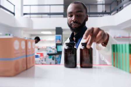 Foto de Farmacéutico afroamericano poniendo pastillas botellas en el estante de la farmacia, enfoque selectivo. vendedor de farmacia tomando paquetes de medicamentos de cerca, la venta de suplementos, vista frontal - Imagen libre de derechos
