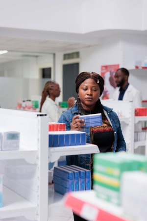 Foto de Mujer afroamericana comprando pastillas en la farmacia, leyendo instrucciones, sosteniendo el paquete de tabletas, de pie cerca del estante de la farmacia. Cliente eligiendo medicamentos, concepto de negocio farmacéutico - Imagen libre de derechos