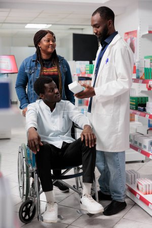 Téléchargez les photos : Consultant en pharmacie afro-américaine aidant l'homme handicapé à choisir des suppléments nutritionnels. Pharmacien recommandant des médicaments au client ayant une déficience physique, toute l'équipe noire - en image libre de droit