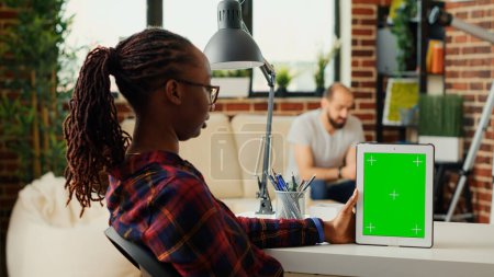 Foto de Freelancer femenina que mira la tableta digital que muestra la pantalla verde en el escritorio de la oficina en casa, utilizando la pantalla de croma key con plantilla de maqueta de pantalla verde. Analizar el espacio de copia aislado en el dispositivo. - Imagen libre de derechos
