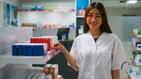 Téléchargez les photos : Travailleur asiatique joyeux examinant les produits pharmaceutiques des étagères de pharmacie, regardant les boîtes à médicaments et les bouteilles de pilules. Pharmacienne vérifiant les vitamines et les emballages de médicaments. - en image libre de droit