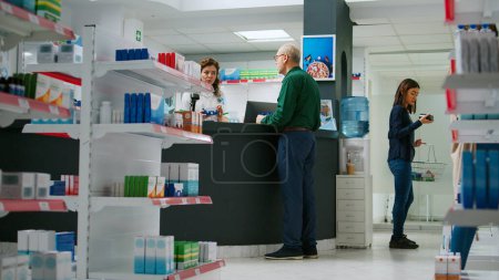 Téléchargez les photos : Client âgé prenant boîte sur les pilules des étagères de la pharmacie pour acheter des médicaments et des soins de santé, parler au pharmacien sur les produits pharmaceutiques. Acheter des suppléments et des médicaments. - en image libre de droit