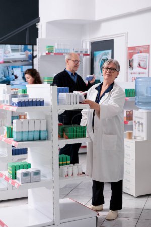 Téléchargez les photos : Pharmacien âgé caucasien debout dans la pharmacie organiser des étagères tout en vérifiant les paquets de pilules, aider les clients avec la prescription de soins de santé. Service et soutien en médecine - en image libre de droit