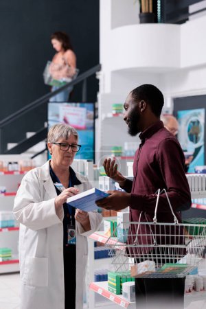 Photo for African american client with shopping basket asking for pharmacist support before buying pills to help cure disease, medicine service. Drugstore employee explaining drugs leaflet working in pharmacy - Royalty Free Image
