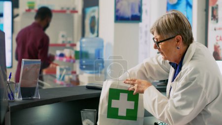 Photo for Senior health specialist putting medicaments and vitamins in pharmaceutical shopping bag, preparing prescription medicine. Elderly woman working at pharmacy cash register counter desk. - Royalty Free Image