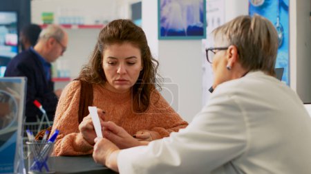 Photo for Woman customer showing prescription paper to pharmacist, asking about disease treatment and medicaments. Young adult talking about box of pills, supplements at cash register. Tripod shot. - Royalty Free Image