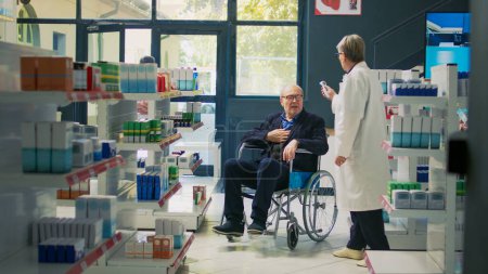Photo for Health specialist measuring temperature on man with impairment, using thermometer with person in wheelchair. Male client buying prescription medicine in disability friendly pharmacy. - Royalty Free Image