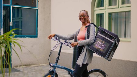 Photo for African american female carrier waiting to customer outdoors, standing next to bicycle to deliver restaurant meal order in thermal bag. Carrying backpack and holding fast food, express services. - Royalty Free Image