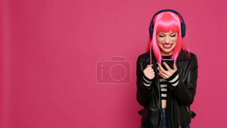 Photo for Cool trendy person listening to music and using social media on smartphone, enjoying songs while she sends messages. Feeling positive and having fun with audio, entertainment over pink background. - Royalty Free Image