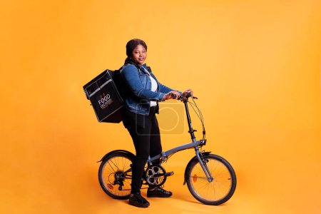 Téléchargez les photos : Femme afro-américaine portant livraison de nourriture sac à dos thermique debout vélo d'équitation en studio avec fond jaune. Employé du restaurant livrant la pizza pour le déjeuner. Service de transport alimentaire - en image libre de droit