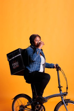 Photo for Portrait of african american deliverywoman sitting on bike while drinking coffee before start delivering takeaway orders to clients during lunch time. Take out food service and concept - Royalty Free Image