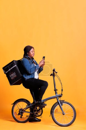 Téléchargez les photos : Employé du restaurant debout sur le vélo en studio avec fond jaune tout en vérifiant adreess client au téléphone avant de commencer la livraison repas à emporter. Pizzeria courier portant sac à dos à emporter - en image libre de droit