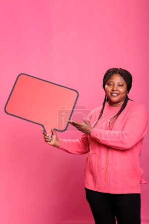 Photo for Joyful smiling adult woman holding blank template announcement placard posing in studio. African american confident woman holding red cardboard speech bubble on pink background. - Royalty Free Image