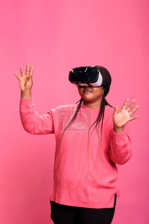 Photo for Cheerful happy woman wearing virtual reality headset playing video games enjoying 3d entertainment during lesiure time. Happy cheerful person with vr headset while standing over pink background - Royalty Free Image