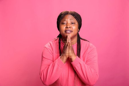 Foto de Retrato del pacífico modelo afroamericano orando a Dios pidiendo perdón mientras estaba de pie en el estudio con antecedentes aislados. Mujer religiosa sosteniendo palmas juntas en oración. Concepto de religión - Imagen libre de derechos