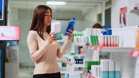 Téléchargez les photos : Jeune femme lisant une notice pharmaceutique en pharmacie, vérifiant les médicaments pour acheter des produits de santé. Client regardant les boîtes de pilules pour acheter un traitement sur ordonnance ou des médicaments. - en image libre de droit