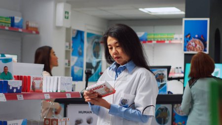 Photo for Asian specialist analyzing boxes of supplements on shelves, working in drugstore and helping sick people. Medical employee checking packages of healthcare pills, pharmacy shop. Handheld shot. - Royalty Free Image