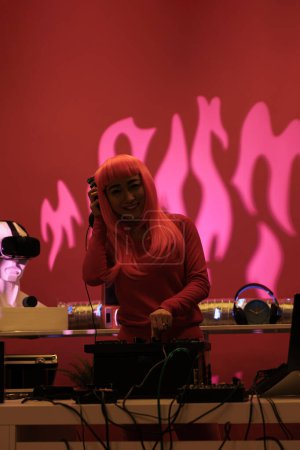 Téléchargez les photos : Asian musical artist working as dj playing at turntables, mixing techno music with eletronic using audio equipment. Performer with pink hair having fun in studio standing over pink background. - en image libre de droit