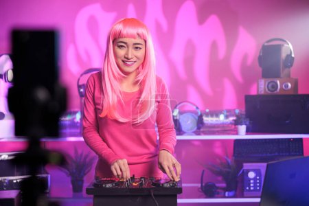 Photo for Asian dj performing techno song using mixer console while recording process with smartphone camera, putting music video on her channel for subscribers. Artist mixing at night in club - Royalty Free Image