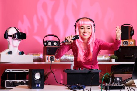 Foto de Asian musician sitting at dj table performing techno music using professional turntables, wearing headset and talking with fans using microphone. Artist with pink hair playing electronic song at night - Imagen libre de derechos