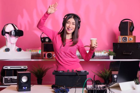 Photo for Asian performer holding cup of coffee while dancing in studio over pink background, playing electronic sound at professional mixer console. Artist doing performance at nightclub with audio equipment - Royalty Free Image