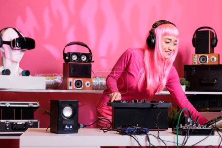 Foto de Asian musician standing at dj table wearing headphones while playing techno music at professional turntables in club at night time. Artist with pink hair having fun while enjoying performing song - Imagen libre de derechos