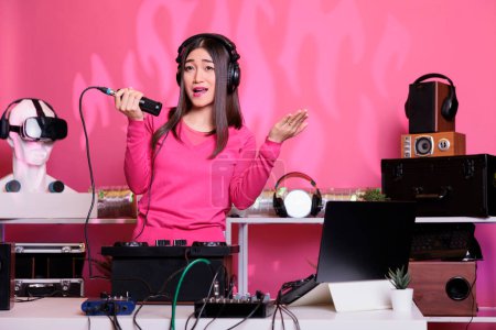 Foto de Smiling performer standing at dj table playing techno music at professional turntables in studio over pink background. Asian musician singing with fans using microphone, having fun during nightlife - Imagen libre de derechos