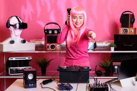 Foto de Asian dj listening techno music at headphones while mixing song at professional mixer console, having fun with fans in club at night. Artist with pink hair doing performance with audio equipment - Imagen libre de derechos