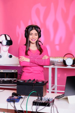 Foto de Portrait of cheerful performer standing with arm crossed in studio over pink background, playing techno music using professional mixer console. Artist mixing sounds, having fun in club at night - Imagen libre de derechos