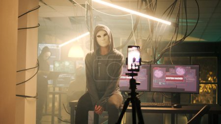 Téléchargez les photos : Diverse team of criminals hiding identity while filming threat video and asking for ransom, leaking valuable information on dark web. Hackers with masks broadcasting live montage. - en image libre de droit